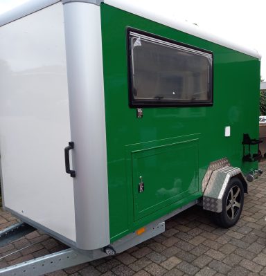 Caravan op maat; fietsendrager of kast Groene aanhanger met een grijze achterkant en een klein raam, geparkeerd op een stenen pad.