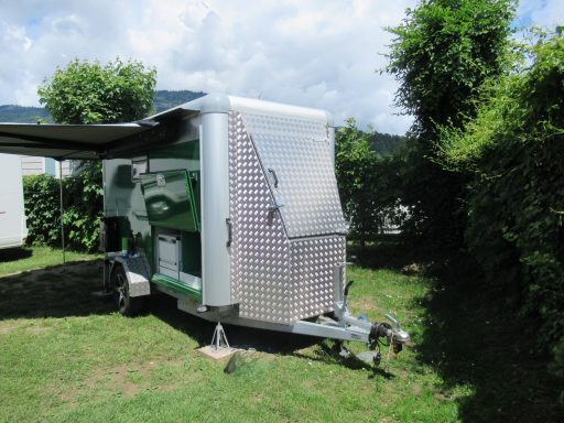 Bergkast voor reservewiel en tuinmeubels Een groene caravan met grote metalen bergkast voor tuinmeubels en uitgedraaide luifel op het gras.