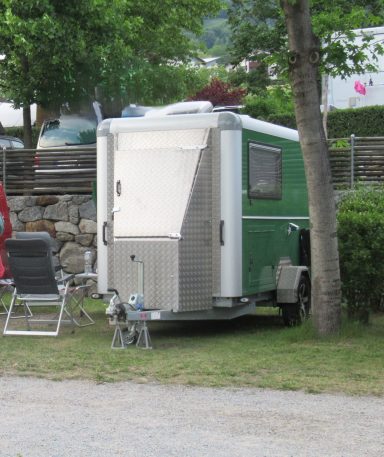 Voorkant caravan op maat Groene caravan met metalen kast op de dissel, naast een stoel en bomen in de omgeving.