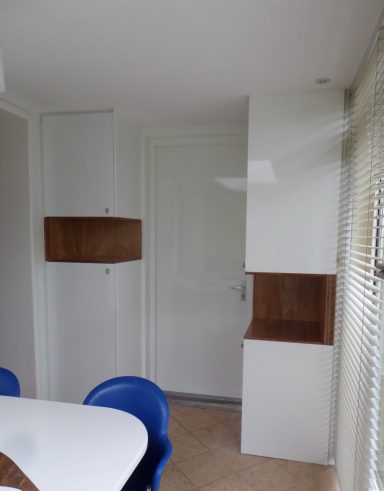 Witte inbouwkasten met houten uitsparingen Moderne ruimte met witte wandkasten met houten accenten en uitsparingen en witte eettafel met blauwe stoelen.