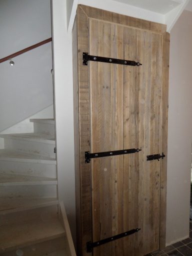 Houten kasteeldeur op maat Houten kasteeldeur met zwarte scharnieren, geplaatst bij een trap.