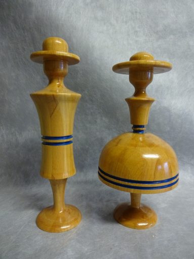 Houten dame en heer sculptuur Twee houten sculpturen, een dame en heer, met hoeden en blauwe strepen op een lichtgekleurde achtergrond.